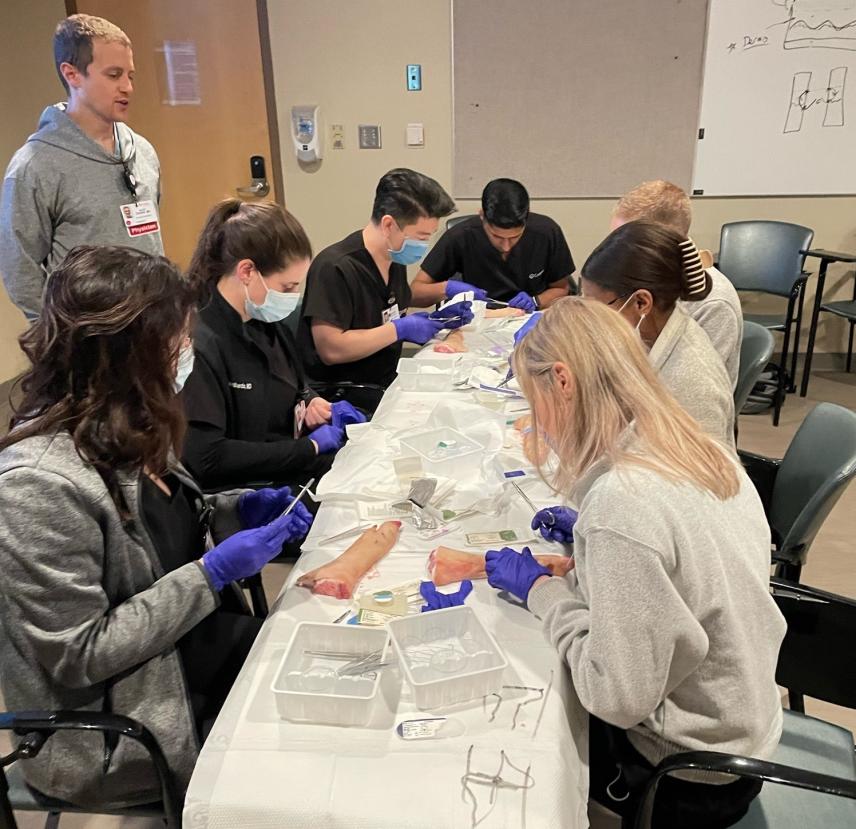Medical residents practicing suturing techniques on training models during a procedure lab, guided by an instructor.